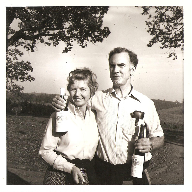 historical black and white photograph of a man and woman with arms around one another, smiling and holding bottles of wine at the edge of a vineyard setting