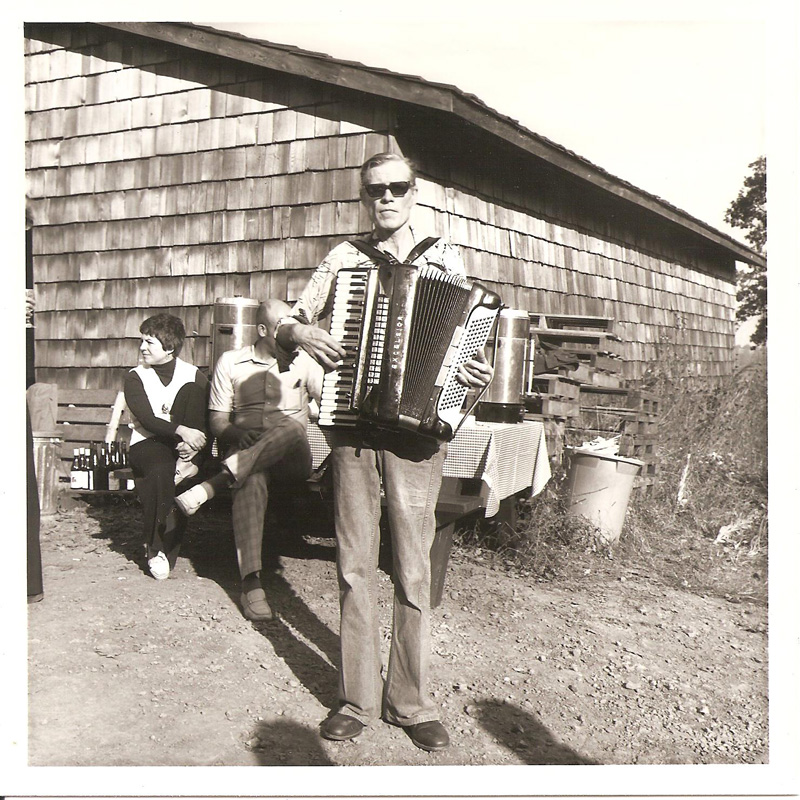 historical black and white photograph of a man standing in front of a cedar shake barn with an accordion