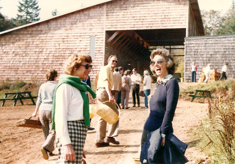 historical photograph of two women smiling widely wearing sunglasses outside a cedar shake barn