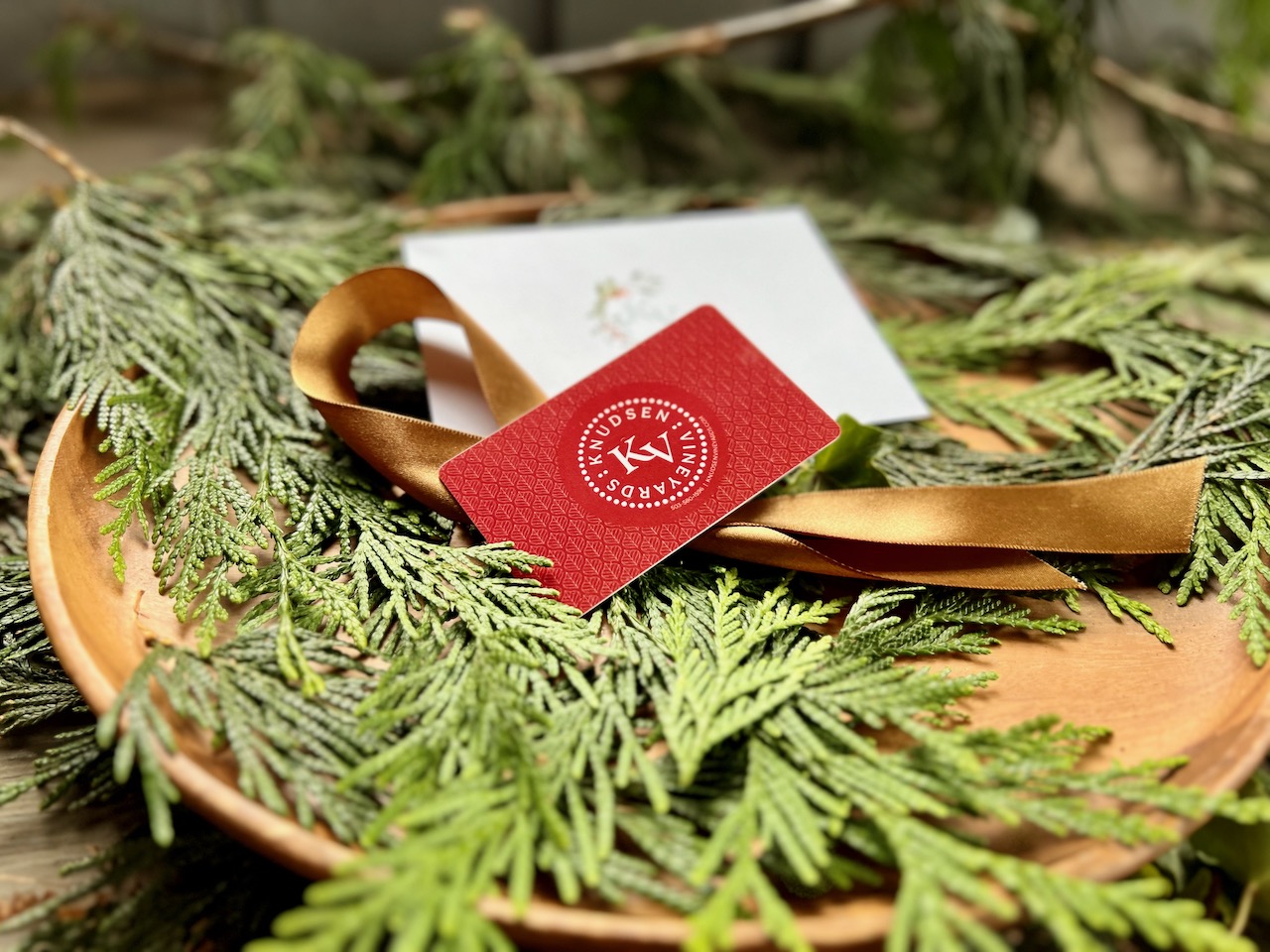 A red gift card with the Knudsen Vineyards logo sits on a lush bed of green pine bows.